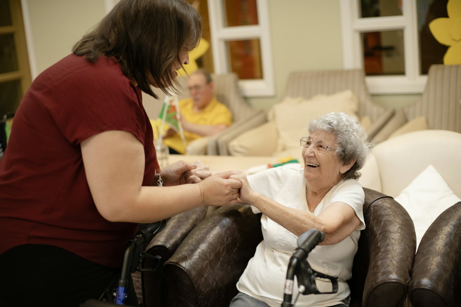 A caregiver holds the hands of a smiling older woman in her living room.
