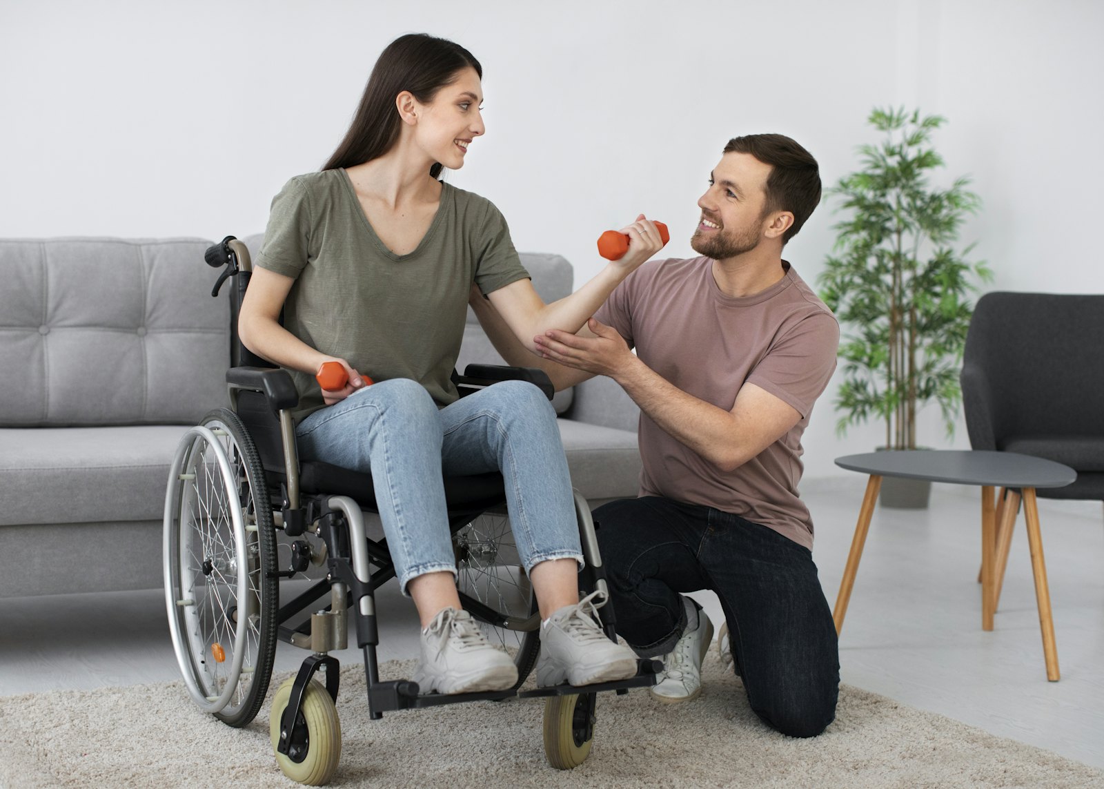 A therapist kneels beside a woman in a wheelchair as she lifts hand weights at home.