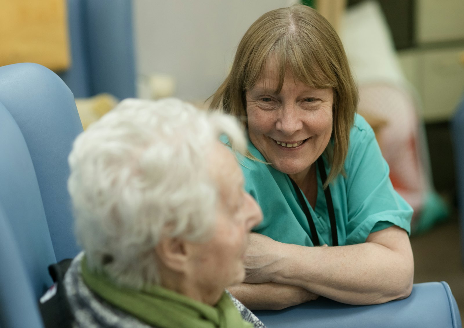 A nurse in scrubs leans in and smiles at an older woman sitting in a chair.